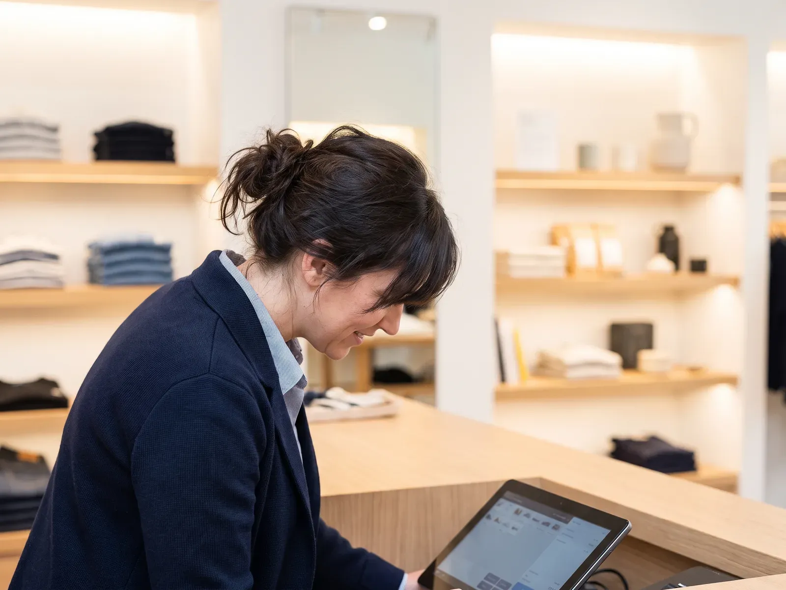 Een vrouw achter een moderne winkelbalie kijkt naar een tablet-kassasysteem, in een lichte boetiek.