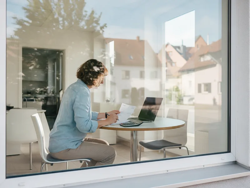Van buiten door het raam gezien: een boekhouder rekent aan een ronde tafel met een rekenmachine, een factuur en een laptop.